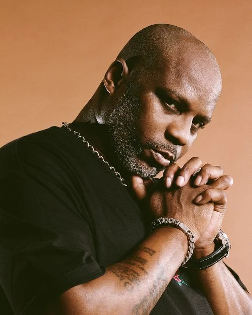 Portrait of African-American man, DMX, with hands together near face, wearing a black t-shirt, chain necklace, and bracelet against tan background.