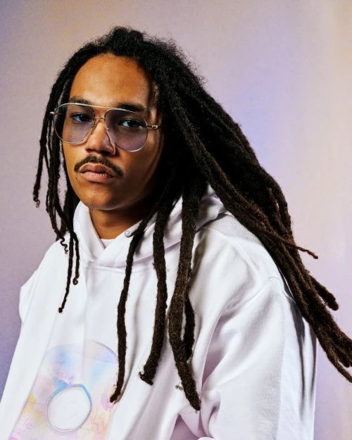 Portrait of an adult male with dreadlocks and glasses wearing a white hoodie, posing against a soft gradient studio backdrop.