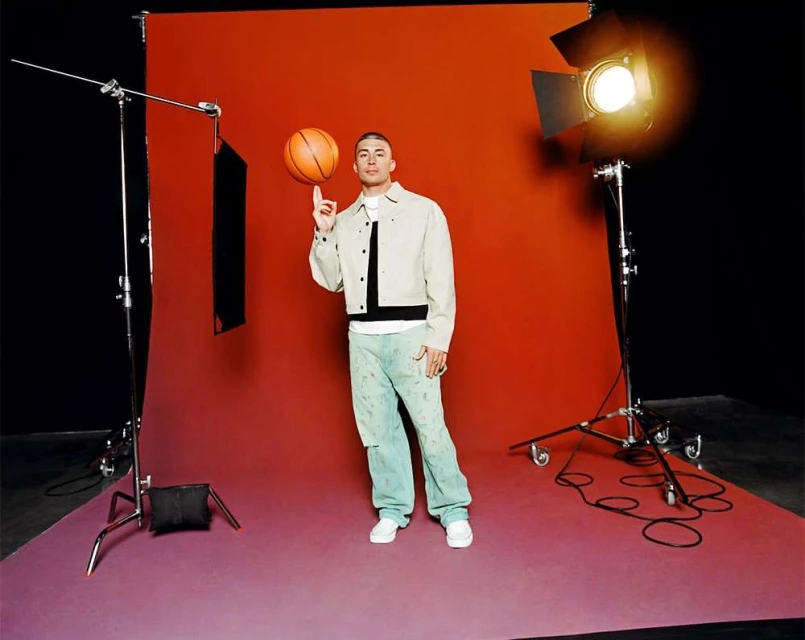An adult male stands centered in a studio setting, skillfully spinning a basketball on his finger, with studio lights visible.