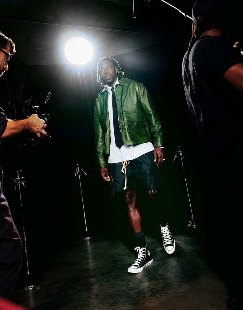 Full-length shot of a Black male model in a leather jacket and shorts, posing in a photo studio with cameras visible on either side.