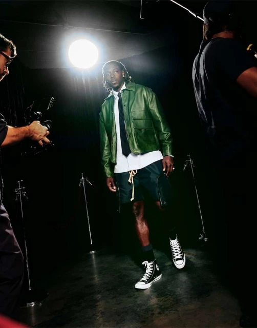 Full-length shot of a Black male model in a leather jacket and shorts, posing in a photo studio with cameras visible on either side.