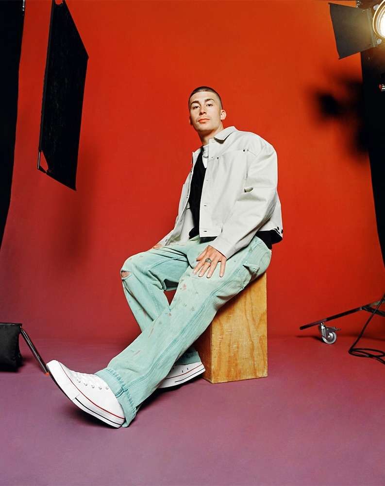 Young adult in white denim jacket and light blue jeans sitting in a photo studio against a solid orange background, wearing Converse shoes.