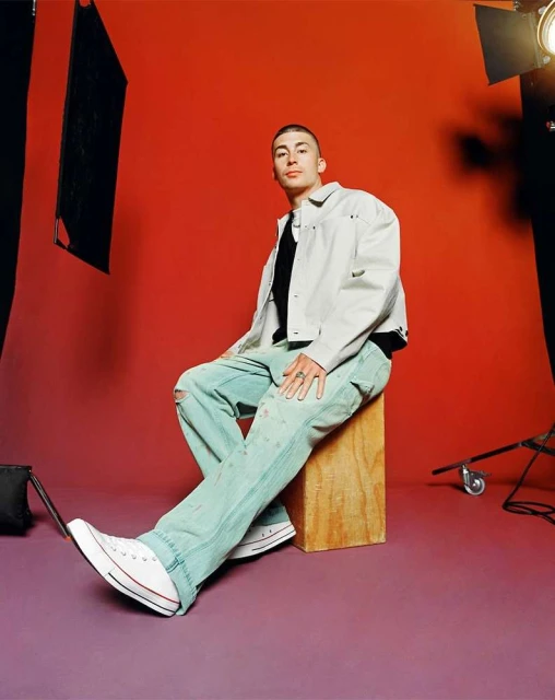 Young adult in white denim jacket and light blue jeans sitting in a photo studio against a solid orange background, wearing Converse shoes.