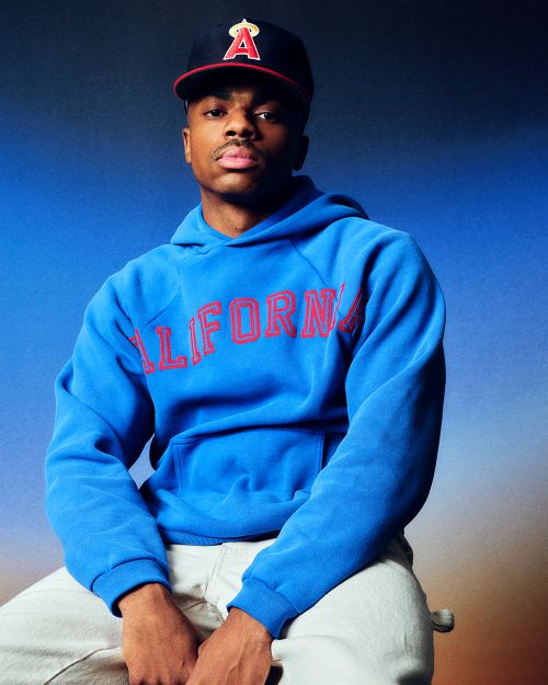 Young adult sits with a blue hoodie and ball cap against a gradient blue background, shot in a studio with soft lighting.