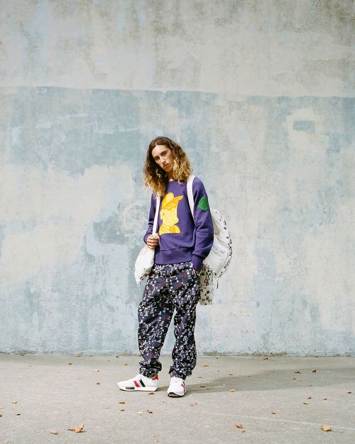 Adult male in patterned pants and purple sweatshirt with cartoon graphic standing against an aged wall, carrying a white backpack.