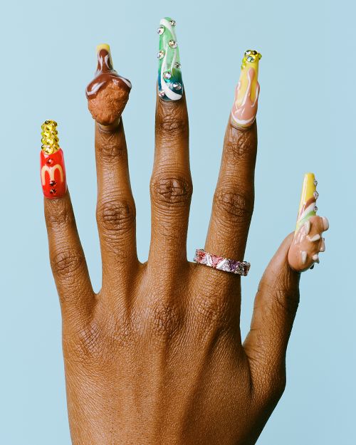 Close-up of an outstretched hand with long decorated fingernails, featuring nail art of fast food items and a sparkling ring.