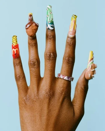 Close-up of an outstretched hand with long decorated fingernails, featuring nail art of fast food items and a sparkling ring.