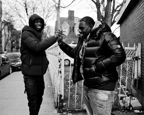 Two young men in puffy jackets greet each other with a handshake on a city street, captured in a high-contrast black and white photograph.