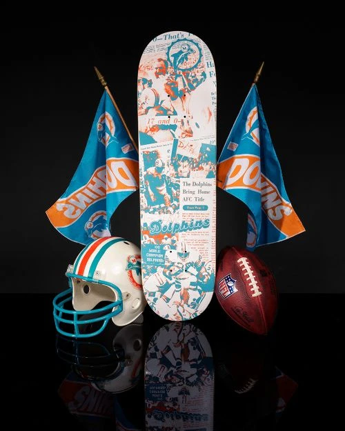 Miami Dolphins football equipment and memorabilia featuring a skateboard, helmet, football, and team flags on a reflective surface.