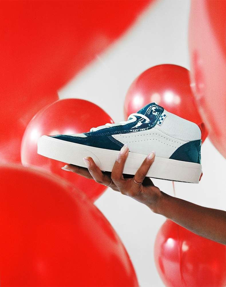 Skateboard shoe in dark blue and white held in hand against a backdrop of red balloons and a white background in a studio setting.