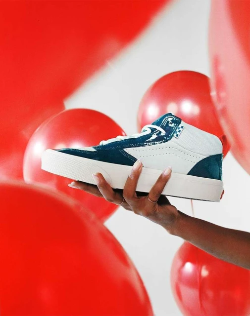 Skateboard shoe in dark blue and white held in hand against a backdrop of red balloons and a white background in a studio setting.