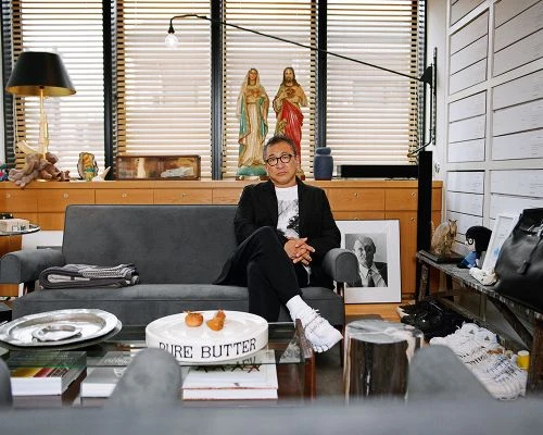 Asian man in a black suit sits on a grey couch in a brightly lit room with religious statues and home decor objects in the background.