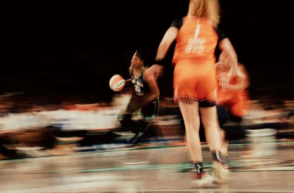 Action shot of two female basketball players in motion on a court during a game; one dribbles the ball, and crowd visible in the background.