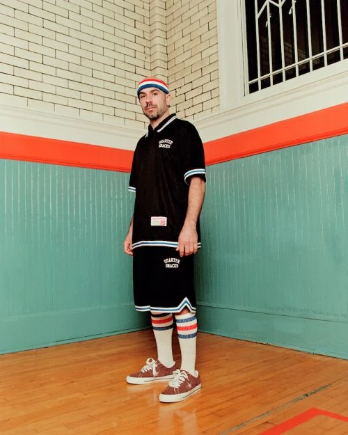Adult male in vintage-inspired athletic wear stands indoors. He wears a striped beanie, black jersey and shorts with sneakers on a wood floor.