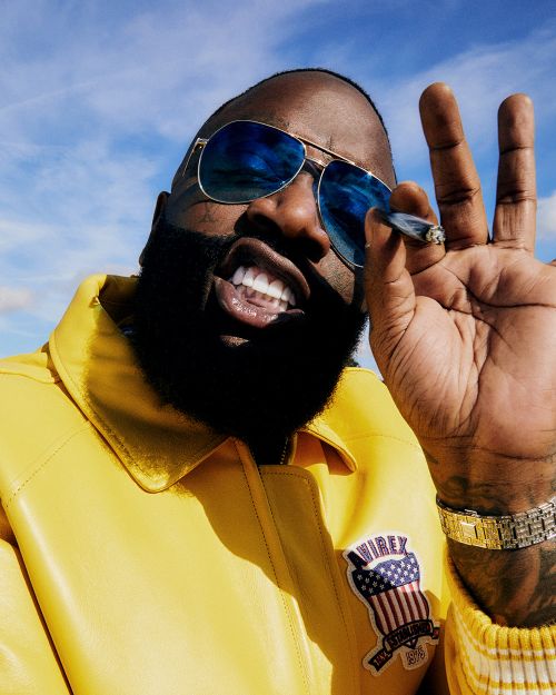 Bearded adult male outdoors wearing aviator sunglasses and a yellow jacket, holding a hand-rolled cigarette in a low-angle portrait. 