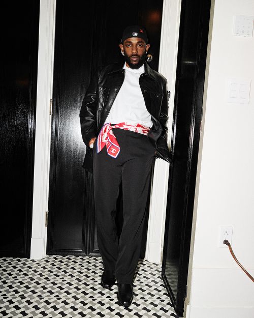 A stylish adult male stands in a black leather jacket and black pants, accessorized with a patterned scarf, indoors with patterned flooring.