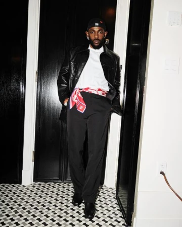 A stylish adult male stands in a black leather jacket and black pants, accessorized with a patterned scarf, indoors with patterned flooring.