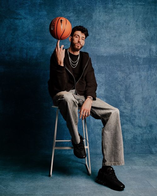 Young adult male seated on a stool, balancing a basketball on his finger in a studio setting with textured blue backdrop.