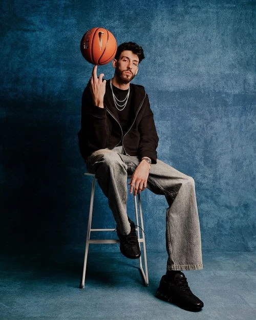 Young adult male seated on a stool, balancing a basketball on his finger in a studio setting with textured blue backdrop.