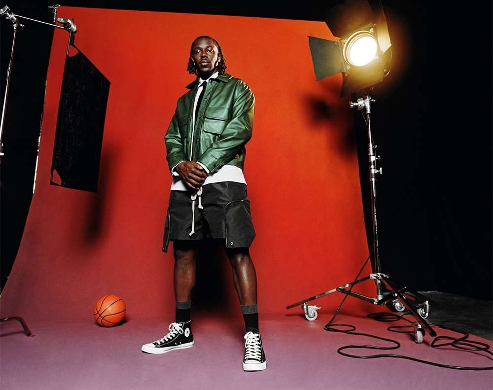 Full shot of a man in a green jacket and shorts, standing in a photo studio with a basketball near lighting equipment.
