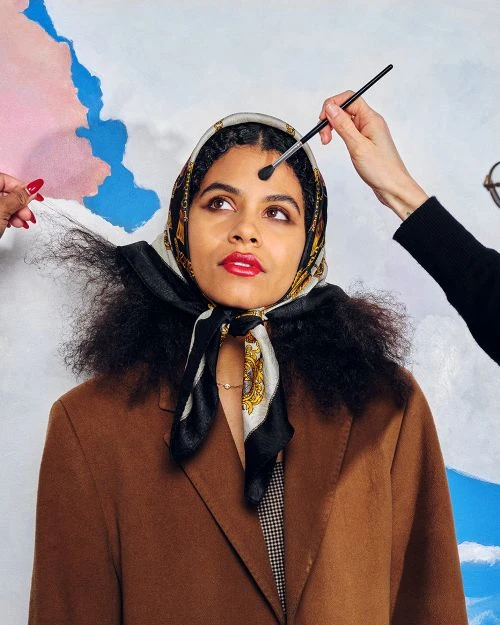 A woman with a headscarf getting makeup applied by several hands against a blue and pink cloud-painted backdrop in a studio setting.