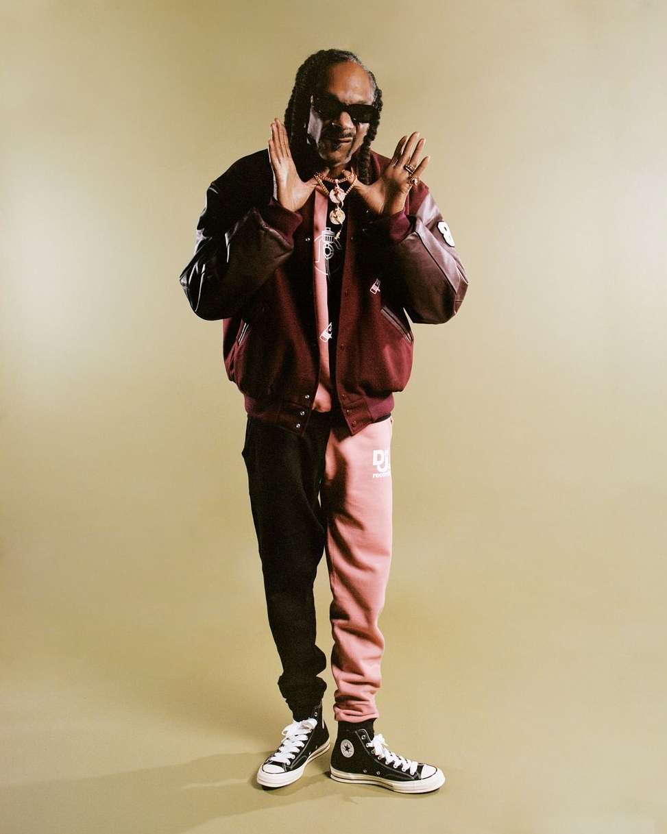 Snoop Dogg stands in a photo studio wearing a burgundy bomber jacket, split-color sweatpants, sunglasses, and black Converse sneakers.