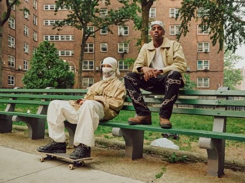 Two people wearing matching tan jackets and head coverings seated on a green park bench in front of a brick apartment building.