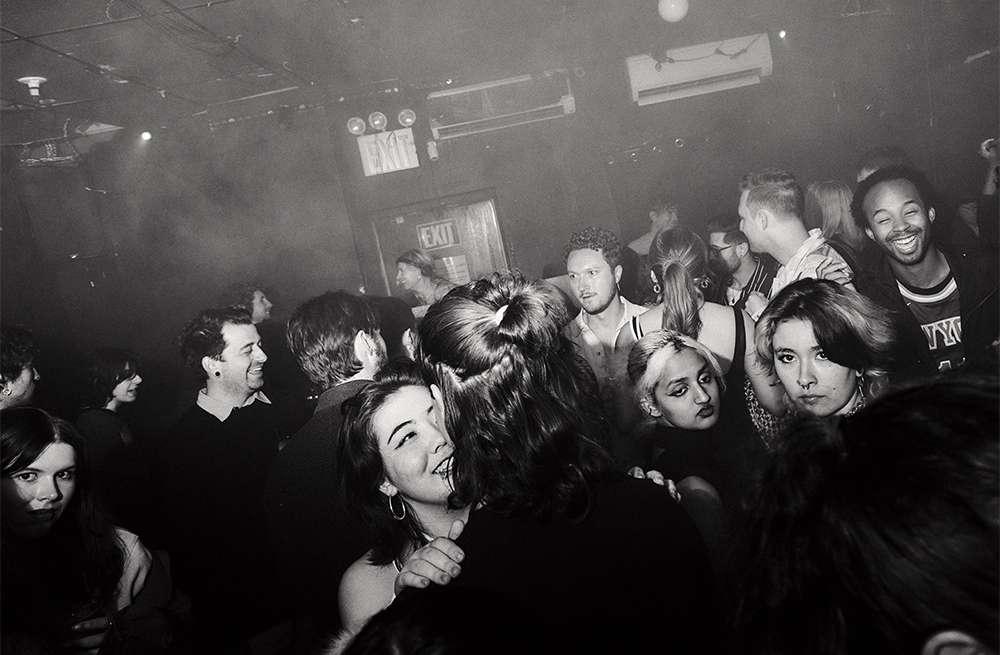 Black and white image captures a lively indoor gathering with a diverse group of people in a dimly lit venue, creating a dynamic scene.