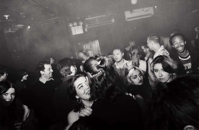 Black and white image captures a lively indoor gathering with a diverse group of people in a dimly lit venue, creating a dynamic scene.