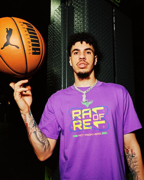 Adult male spins a Puma basketball on his finger. He wears a purple t-shirt and chain necklace indoors against a dark background.