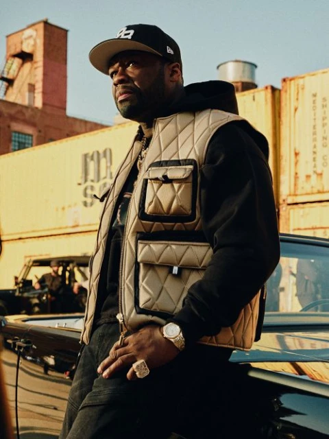 Rapper 50 Cent leans on a black car outdoors wearing a quilted vest, baseball cap, jewelry, and black clothing in an urban environment.