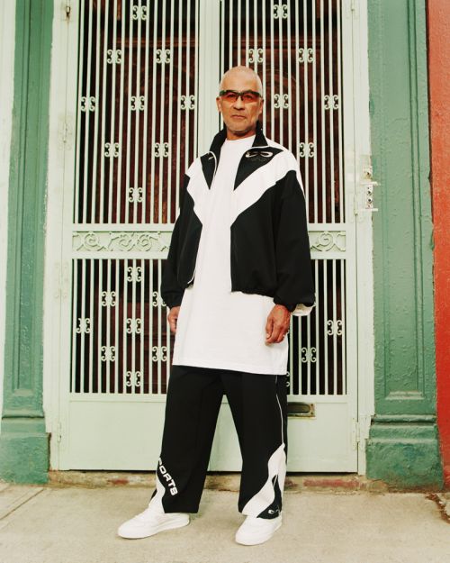 Adult man in a black and white tracksuit stands in front of ornate doors with orange and green walls. Full shot, eye-level angle.