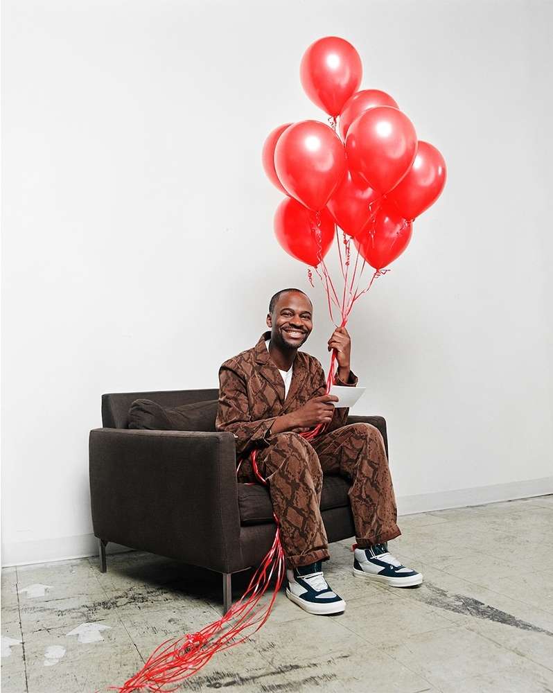 Smiling adult male sitting in a chair holds a bunch of red balloons. He wears a brown pattern jacket and pants in a white room.