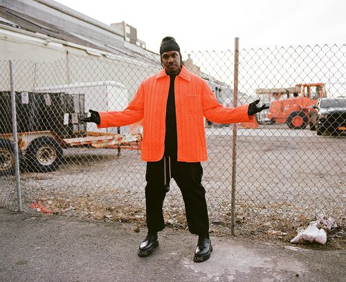 Adult male poses outdoors in an orange jacket, black pants, and black beanie, standing in front of a chain-link fence with a construction vehicle visible.
