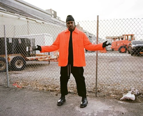 Adult male poses outdoors in an orange jacket, black pants, and black beanie, standing in front of a chain-link fence with a construction vehicle visible.