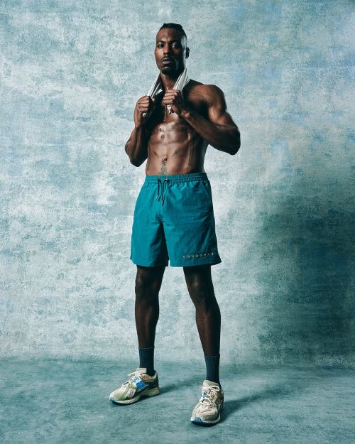 Athletic, shirtless African American man with a towel around his neck stands in workout shorts and sneakers against an aqua background.