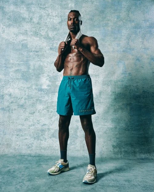Athletic, shirtless African American man with a towel around his neck stands in workout shorts and sneakers against an aqua background.