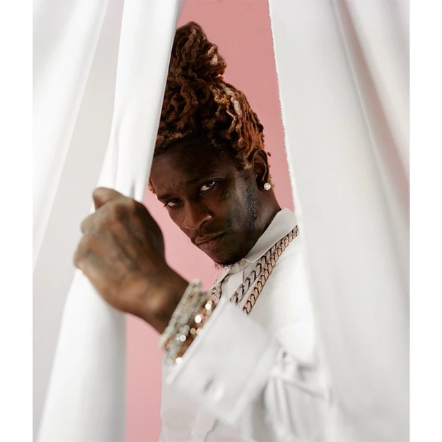 Portrait of a man in a white shirt and gold jewelry behind a curtain. The background is light pink. He has light-brown dreadlocks.