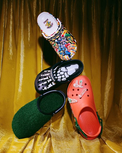 A stack of four Crocs clogs in various colors and designs arranged against a draped gold curtain backdrop, product-style photography.