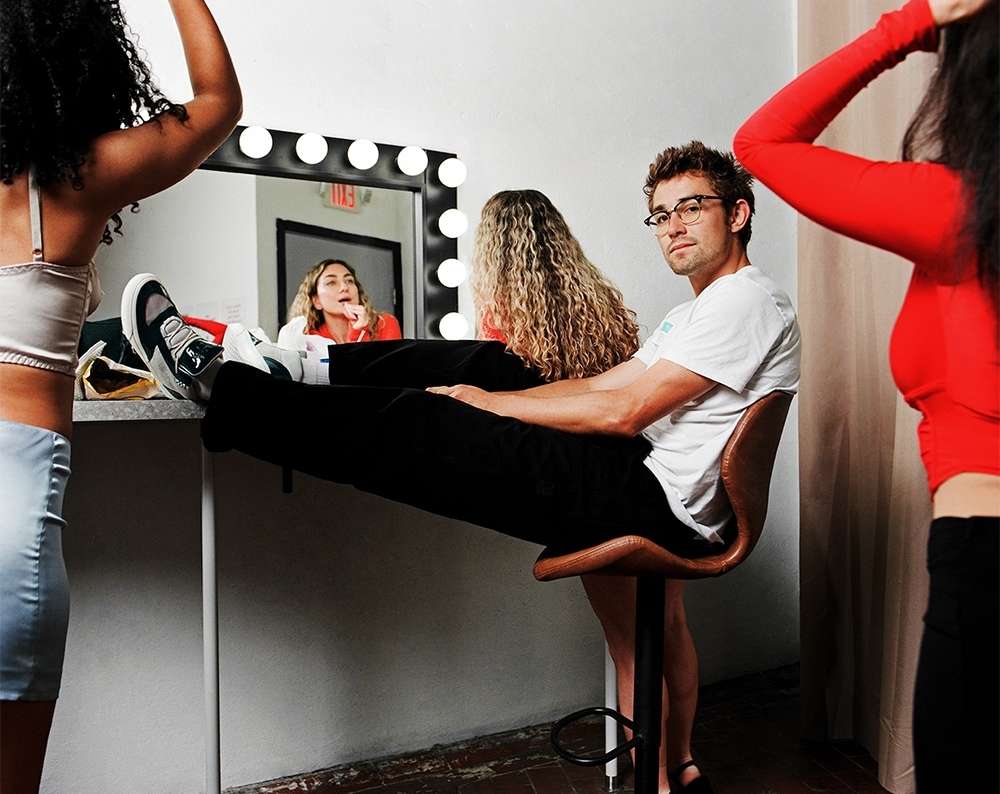 A man sits on a stool in front of a makeup mirror, with two women in the background and foreground of an indoor setting.