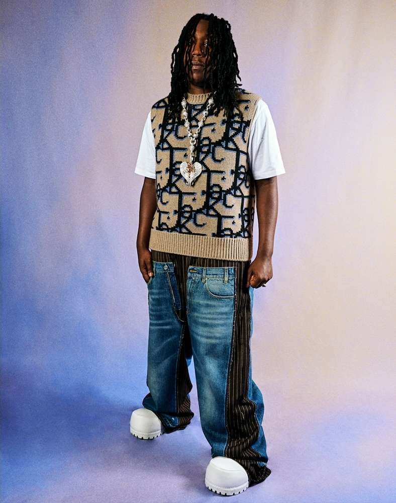 Full-length shot of a Black male wearing oversized, pieced blue jeans and a patterned vest with white platform shoes in a studio setting.