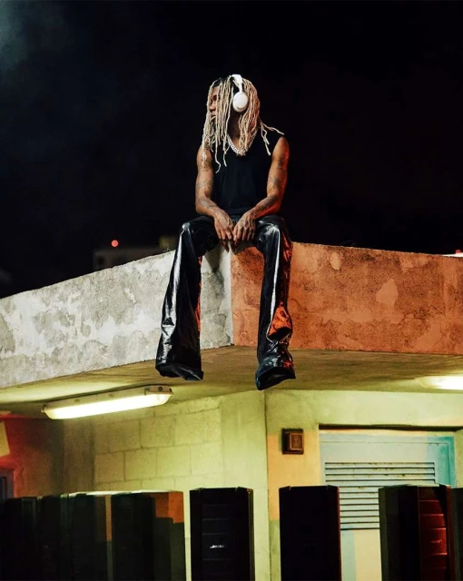 Man with dreadlocks and headphones sitting on a rooftop at night, wearing shiny pants and a sleeveless black shirt, with lighting below.