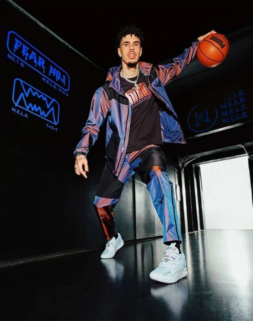 Athletic male model in an iridescent track suit and white sneakers holding a basketball in a dark studio with neon sign.