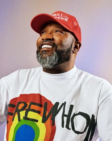 Close-up shot of an adult male with a beard wearing a red cap and a white t-shirt with a rainbow design smiling and looking upwards.