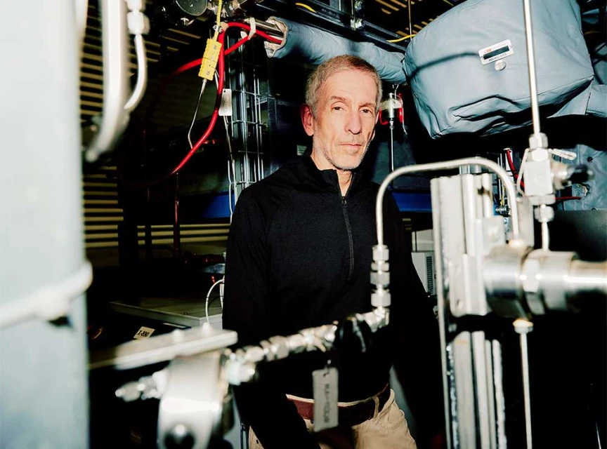 Indoor portrait of a mature adult male in a dark jacket, looking toward the camera, surrounded by machinery and industrial equipment.