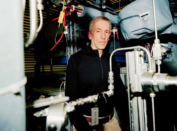 Indoor portrait of a mature adult male in a dark jacket, looking toward the camera, surrounded by machinery and industrial equipment.