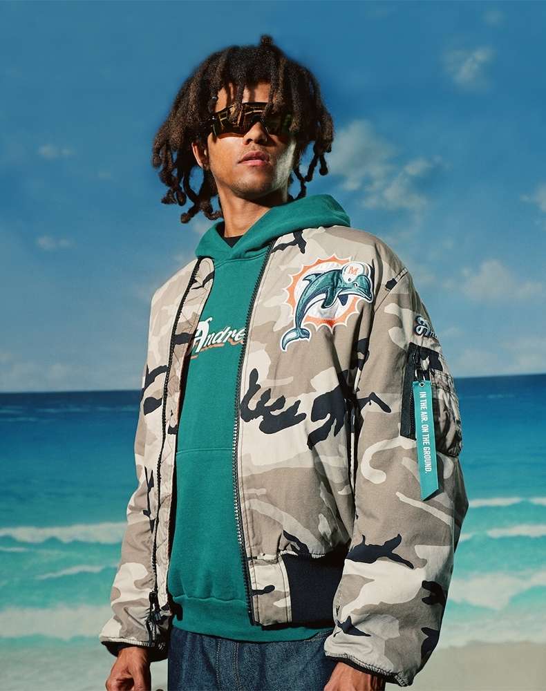 Man in camouflage jacket with Miami Dolphins logo, green hoodie, and sunglasses stands on a beach with blue sky and ocean.