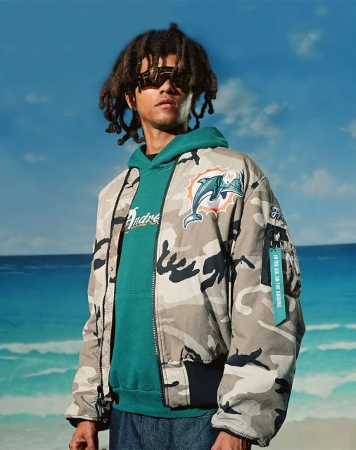 Man in camouflage jacket with Miami Dolphins logo, green hoodie, and sunglasses stands on a beach with blue sky and ocean.