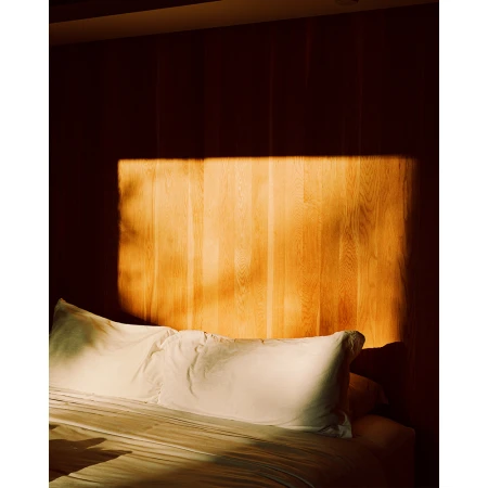 Bedroom interior with white bed and warm wood-paneled wall, illuminated by soft, diffused light creating a cozy, inviting atmosphere.