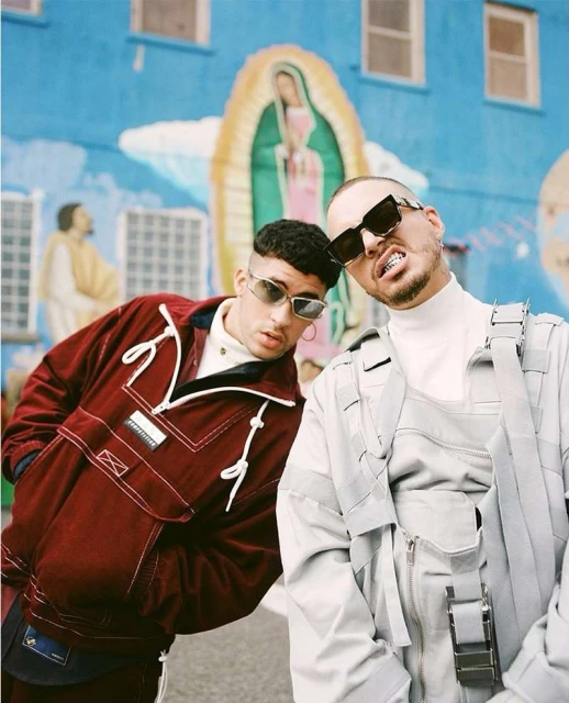 Two musicians, Bad Bunny and J Balvin, pose outdoors in front of a colorful mural. They wear fashionable clothing and sunglasses.